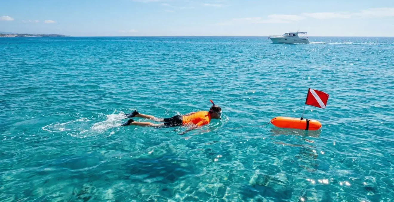 Randonneur palmé utilisant une bouée de signalisation orange vif dans une eau turquoise avec un bateau à moteur visible au loin