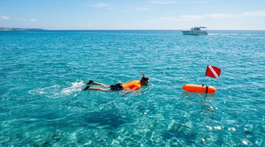 Randonneur palmé utilisant une bouée de signalisation orange vif dans une eau turquoise avec un bateau à moteur visible au loin